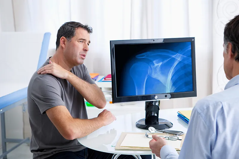 Representative image for acromioclavicular joint dislocation - The image shows a man experiencing pain and discomfort in his shoulder during a medical consultation.